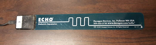 A blue-green, rectangular Decagon Devices ECH20 sensor sitting on a wood surface.