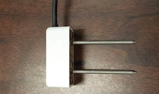 A T shaped, white, Decagon Devices GS1 sensor sitting on a wood surface.