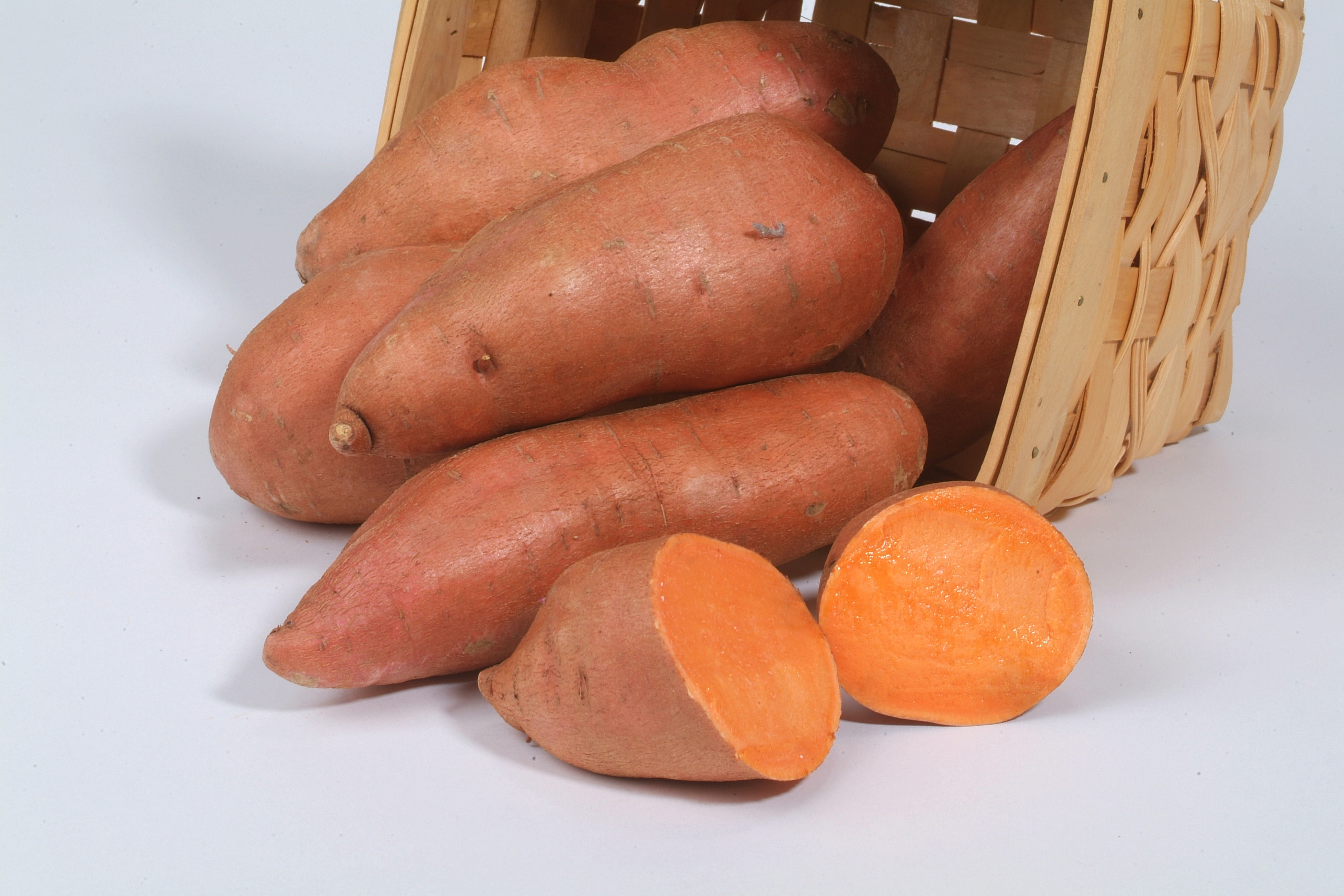 Sweet potatoes with orange skin in basket with one cut open revealing deep orange flesh.