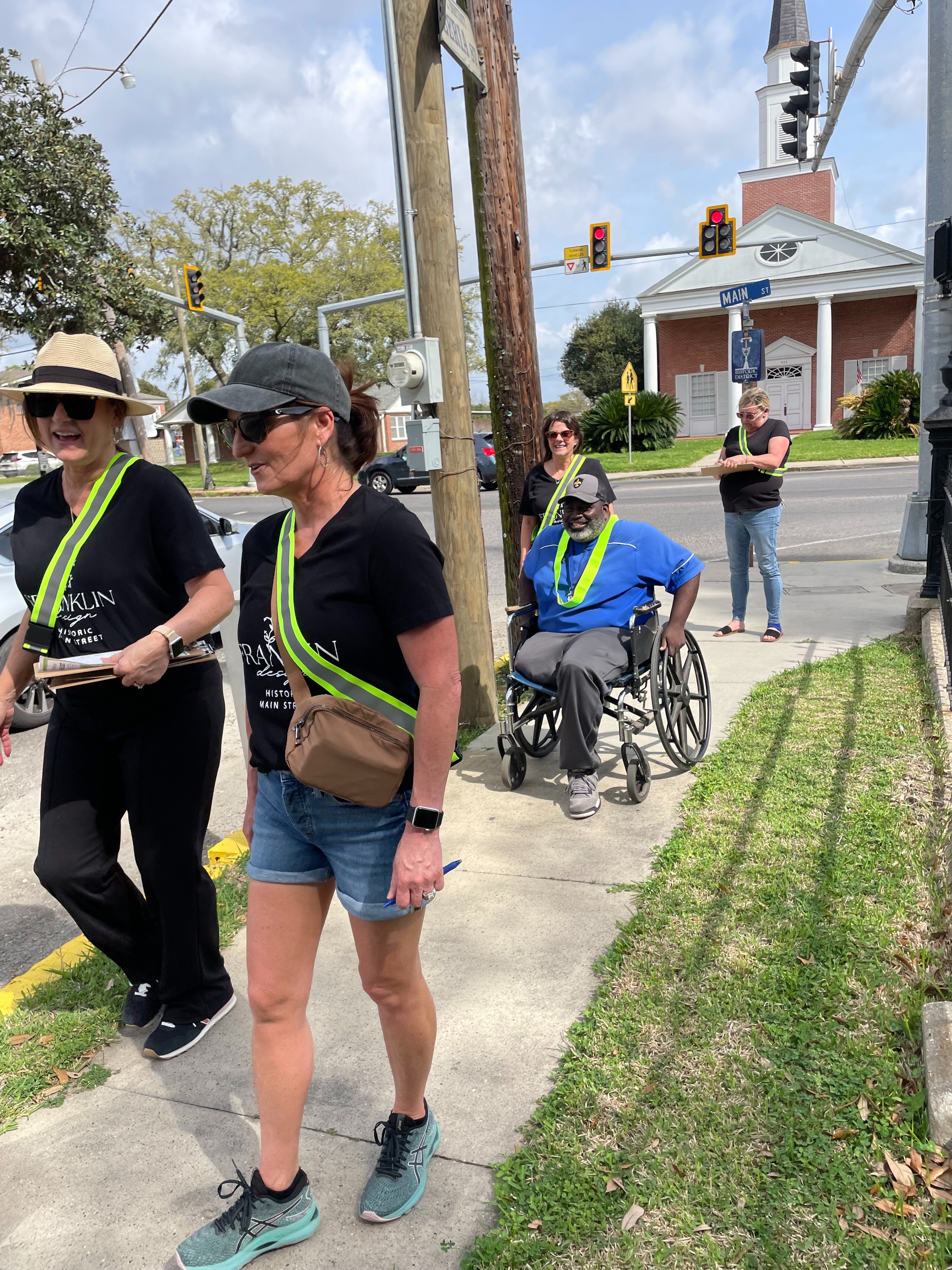 Two women wearing florescent yellow sashes walk on a sidewalk ahead of a man in a wheelchair and two more women. 