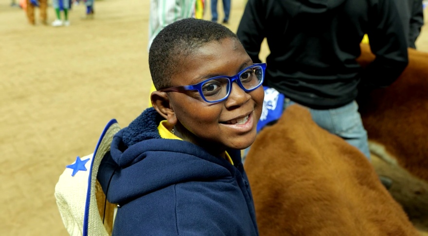 Child wearing a backpack stands beside a brown calf in an indoor livestock arena.
