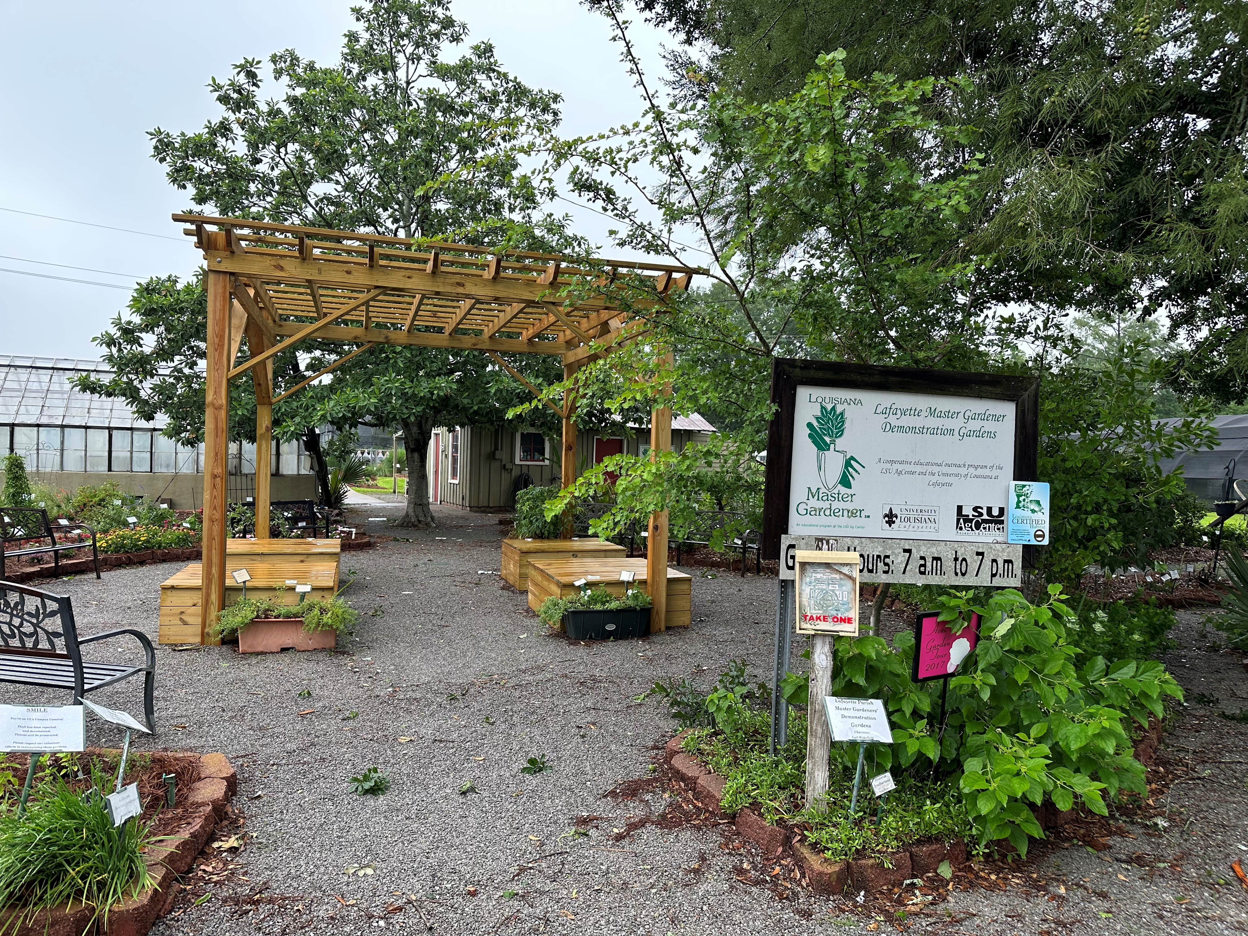 Wooden pergola with seating around the edge. Trees and plants surround two sides of the pergola. Raised demonstration bed gardens are on the side of the pergola, with the front being open.