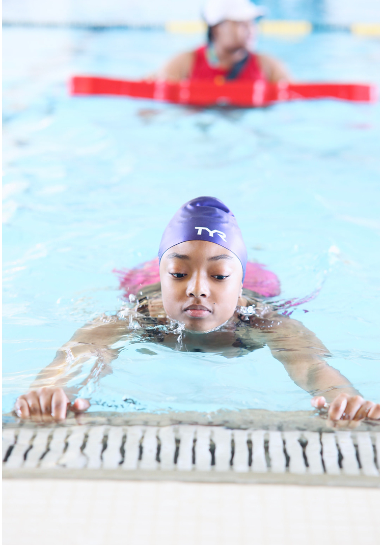 A girl wearing a swim cap floats while hanging on to the side of the pool during a swimming lesson. 
