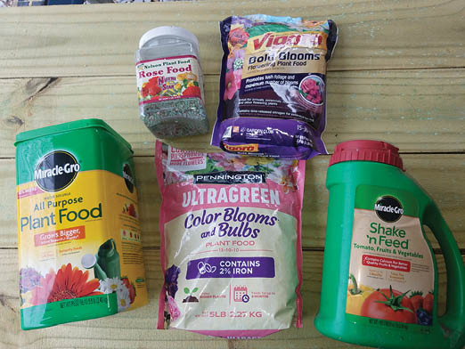 A photo of five containers of different brands of slow-release and water-soluble fertilizers arranged on a table in two rows. The top row is a clear plastic square container with Nelson Plant Food Rose Food and a bag of Vigoro Bold Blossoms Flowering Plant Food. The bottom row is a square, gree plastic container of Miracle-Gro Water Soluble All Purpose Plant Food, a 5 pound bag of Pennington Ultragreen Color Blooms and Bulbs Plant Food 15-10-10, and the last one Miracle Gro Shake 'n Feed Tomato, Fruits & Vegetables in a large green plastic container with red lid.